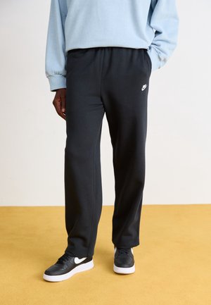 Personne portant un pantalon de jogging noir Nike et des baskets noires Nike, debout sur un sol jaune avec un haut à manches longues bleu clair.