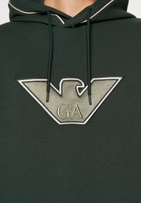 Mörkgrön huvtröja i mjukt material med en stor, broderad örnlogotyp i ljusgrått och "GA" under. Dragsnörena är mörkgröna.