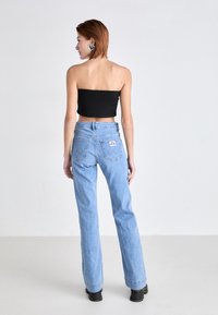 Femme aux cheveux rouges courts portant de grandes boucles d'oreilles en forme de cœur, un top noir sans bretelles, un jean bleu clair taille haute et des bottes noires, dos tourné.