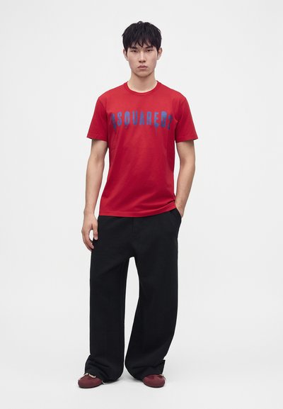 Νέος άνδρας που στέκεται φορώντας κόκκινο t-shirt Dsquared2, μαύρο παντελόνι με φαρδιά πόδια και μπορντό παπούτσια σε απλό λευκό φόντο.