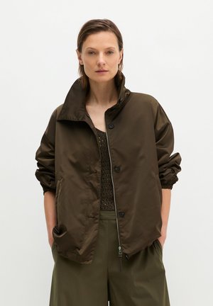 Stehende Frau trägt eine lockere dunkelbraune Jacke mit Reißverschluss, passende Hose und ein strukturiertes dunkelbraunes Top vor schlichtem weißem Hintergrund.