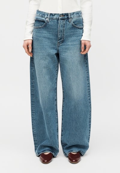 Jean en denim bleu à jambes droites porté avec une chemise blanche à manches longues et des chaussures bordeaux à bout pointu.