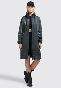 Szürke, fényes, térdközépig érő esőkabát cipzáros zsebekkel, húzózsinóros kapucnival és rugalmas mandzsettákkal, fekete crop top-pal és térd fölé érő fekete csizmával párosítva.