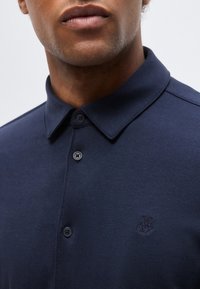 Άνδρας που φοράει μπλε ναυτικό πόλο με κουμπιά, γιακά και μικρό κεντημένο λογότυπο "MOP" στο αριστερό στήθος.