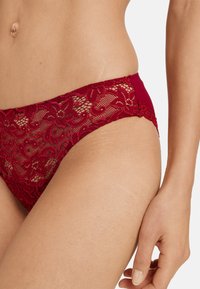 Bikini in pizzo di un rosso scuro con motivo floreale e bordi ondulati, caratterizzato da una fascia in tessuto liscio per il comfort e un taglio seducente.