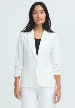 Pikkade tumejuustega naine, kes kannab valget jopet musta pealepaela kohal ja sobivaid valgeid pükse, seistes ühevärvilise hele tausta ees.