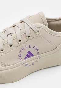 Béžové plátěné tenisky s kulatým fialovým logem, sladěnými tkaničkami a gumovou podrážkou; mají zesílené očka a strukturovaný povrch.