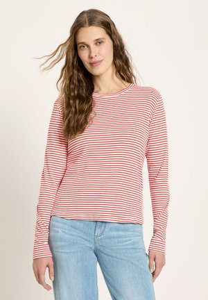 Woman with long wavy brown hair wearing a red and white striped long-sleeve shirt and light blue jeans, standing against a plain background.