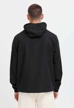 Mann, von hinten gesehen, trägt eine schwarze Kapuzenjacke und hellfarbene Hose, steht vor einem einfachen hellgrauen Hintergrund.