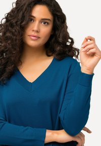 Türkiser V-Ausschnitt-Pullover mit gerippten Bündchen und Saum. Weiche Stricktextur, lockere Passform, die eine natürliche Silhouette betont. Das Model gestikuliert mit der Hand.
