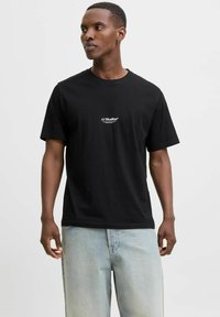 Camiseta de algodón negra con mangas cortas, cuello redondo y pequeño logo blanco "JJ Studios" en el pecho; combinada con jeans de mezclilla azul claro.