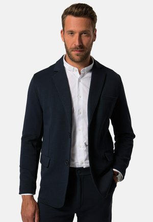 Marineblazer mit Reverskragen, einem Knopfverschluss und zwei Vordertaschen. Getragen über einem weißen Hemd mit Knöpfen. Glatte Textur.