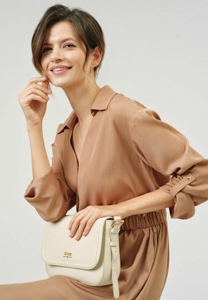 Kremowa torebka crossbody z ekoskóry, z klapą i regulowanym paskiem, umieszczona na modelce w beżowej satynowej bluzce.
