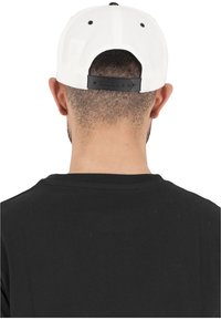 Cappellino da baseball bianco con una striscia nera regolabile sul retro, visto da dietro. Sotto il cappello si intravede una maglietta nera.