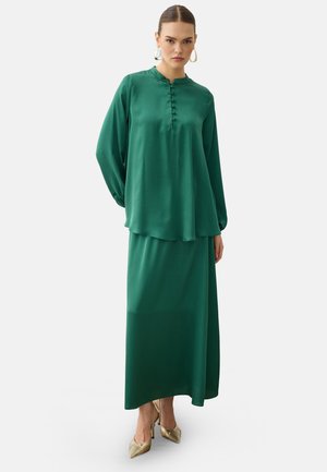 Mujer con blusa de satén verde oscuro de manga larga y falda larga a juego, complementada con grandes pendientes y zapatos de tacón dorados.