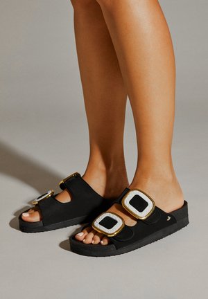Sandales noires avec une épaisse semelle, deux boucles surdimensionnées en or et noir, fabriquées en matériau doux, avec un design à bout ouvert.