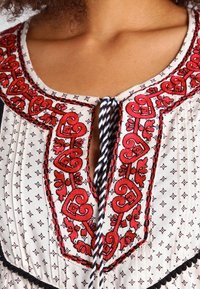 Vit blus med intrikata röda broderier i hjärt- och blommönster. Texturerad tyg med nyckelhålsutskärning och flätad kant.