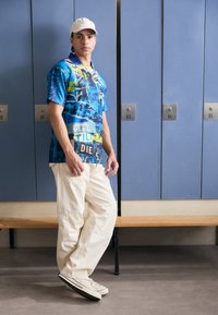 Kleurrijk graphic t-shirt met blauwe, gele en zwarte patronen, gecombineerd met witte, loszittende cargo-broeken en beige sneakers. Licht honkbalpet.