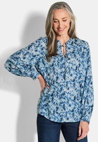 Blaue gemusterte Bluse mit langen Ärmeln, rundem Ausschnitt und einer Schleifendetail vorne. Der Stoff hat eine glatte Textur und einen fließenden Schnitt.