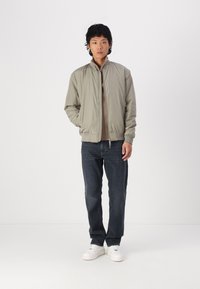 Giacca bomber color beige chiaro con polsini a coste, indossata sopra un top marrone, abbinata a jeans di denim scuri e sneakers bianche. Design semplice e casual.