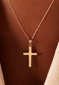 Gouden kruis hanger op een delicate ketting, rustend tegen een gebruinde huid. De hanger heeft een gladde, gepolijste afwerking.