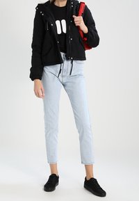 Černá kapucová bunda, černé tričko s grafikou, světle modré džíny s vysokým pasem a černé tenisky. Červená taška držena v jedné ruce. Ležérní outfit.