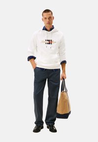 Sudadera blanca con logo, camiseta azul marina debajo, pantalones azul oscuro, zapatos negros, sosteniendo una bolsa beige y azul marina, de pie sobre un fondo liso.