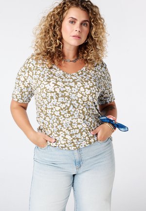 Vrouw met krullend blond haar, die een groen-wit bloemenoverhemd en lichtblauwe spijkerbroek draagt, staand met handen in de zakken tegen een witte achtergrond.