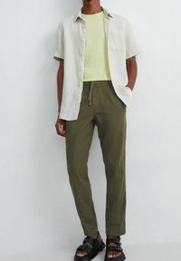 Lniana koszula z krótkim rękawem w jasnobeżowym kolorze, zestawiona z żółtą koszulką i oliwkowozielonymi spodniami; czarne sandały z paskami.