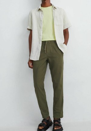 Lněná košile s krátkým rukávem ve světle béžové barvě, doplněná žlutým tričkem a olivově zelenými kalhotami; černé sandály s pásky.