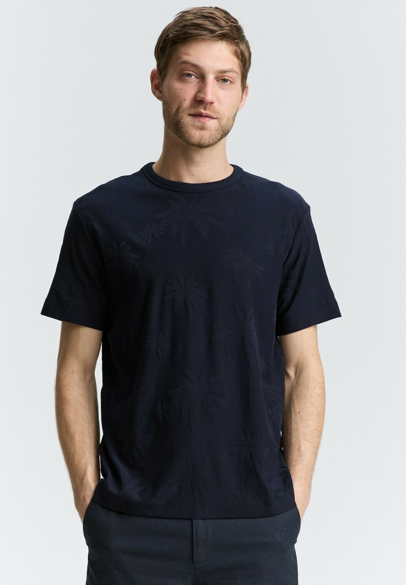 Navyblauwe katoenen t-shirt met een gestructureerd palmbladpatroon. Korte mouwen en een ronde hals. Model staat met handen in de zakken.