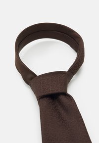 Brown fabric tie featuring a textured pattern, with a smooth finish. The tie is neatly knotted at the top, displaying a wider end.