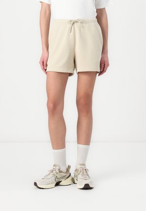 Person iført beige shorts med snørre, hvide mellemlange sokker og beige Nike-sneakers står mod en hvid baggrund.