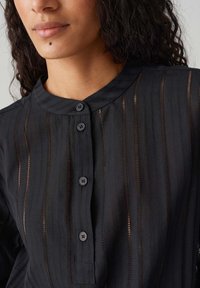 Camicia nera con bottoni, caratterizzata da un colletto rotondo, strisce verticali trasparenti e una delicata trama. Quattro bottoni neri decorano la parte anteriore.