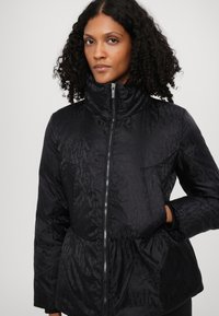 Black puffer jacket with a high collar, featuring a tonal text pattern, zip closure, elasticized waistband, and side pockets. Soft material.
