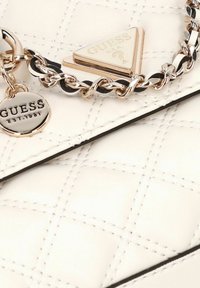 Witte gewatteerde leren tas met gouden kettingriem, voorzien van een ronde hanger en driehoekige logoplaat gegraveerd met "GUESS USA".