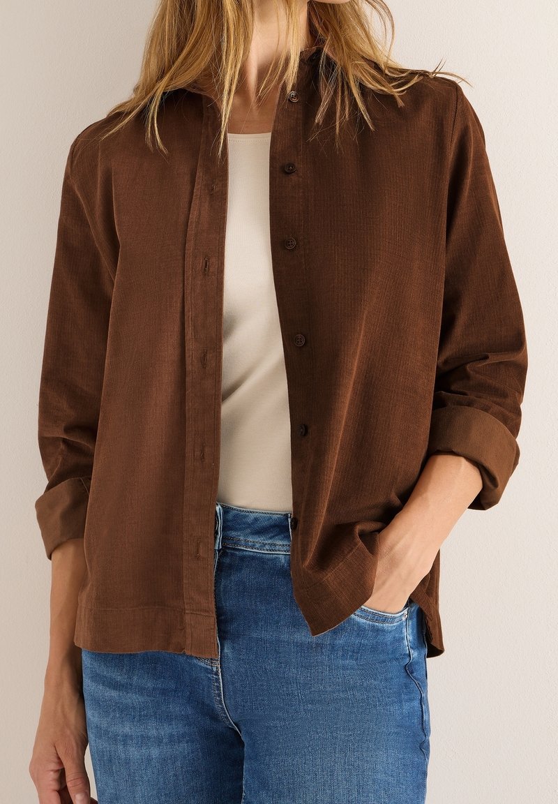 Personne portant une chemise marron à boutons par-dessus un haut beige avec un jean bleu, main dans la poche, debout devant un fond clair uni.