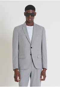 Giacca grigia chiara con revers, due tasche frontali, chiusura a bottone singolo e tessuto texturizzato; indossata sopra una t-shirt bianca.