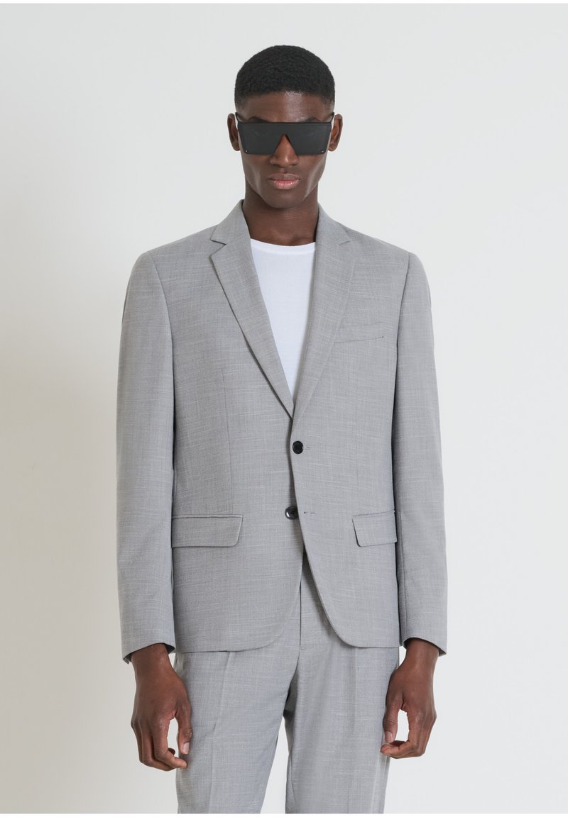 Giacca grigia chiara con revers, due tasche frontali, chiusura a bottone singolo e tessuto texturizzato; indossata sopra una t-shirt bianca.