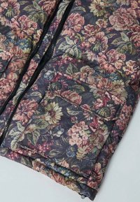 Veste matelassée florale avec un fond sombre, ornée de fleurs roses, jaunes et vertes. Comprend une fermeture éclair et des détails de poche avant.
