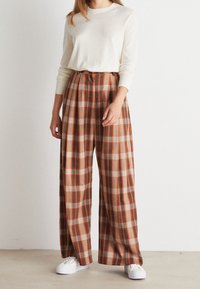 Beige dlouhý rukáv top spárovaný s vysokým páskem, širokými kalhotami s kostkovaným vzorem v oranžové, hnědé a krémové barvě. Bílí tenisky dokončují vzhled.