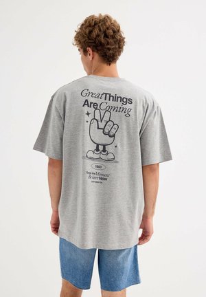 Joven con cabello rizado que lleva una camiseta gris con una mano de dibujo animado mostrando el signo de la paz y el texto "Grandes cosas están por venir", combinado con pantalones cortos azules.