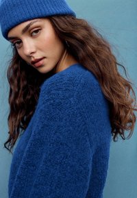 Modrý pletený sveter s jemnou textúrou a voľným strihom, doplnený o zladenú rebrovanú čiapku. Pozadie je jednofarebné svetlomodré.