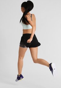 Atletický outfit složený z světle modrého sportovního podprsenky, černých šortek s síťovinovým akcentem a modrých tenisek s bílou podrážkou, v pohybu.