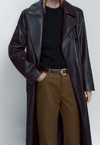 Dark brown leather coat with peak lapels, long sleeves, and a single button, paired with a black sweater and brown trousers with a belt.