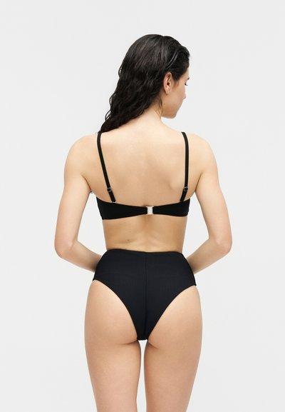 Γυναίκα με μακριά σκούρα μαλλιά που φοράει μαύρο μπικίνι δύο τεμαχίων, εμφανίζεται από πίσω με φόντο ένα απλό φωτεινό υπόβαθρο.
