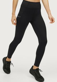 Zwarte high-waisted leggings van rekbaar materiaal, met een zijzak en naaddetails. Gecombineerd met zwarte sport schoenen.