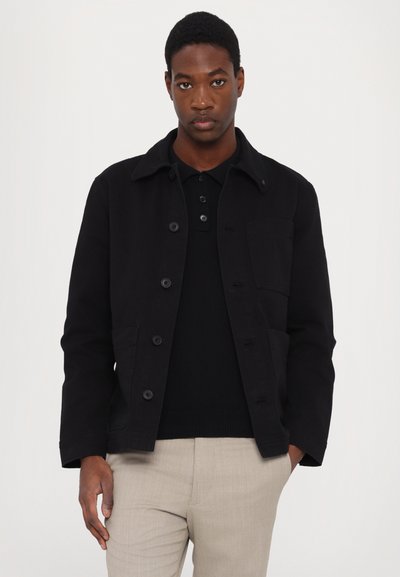 Black cotton jacket with a collar, front buttons, and chest pocket. Worn over a black polo shirt, paired with light-colored trousers.