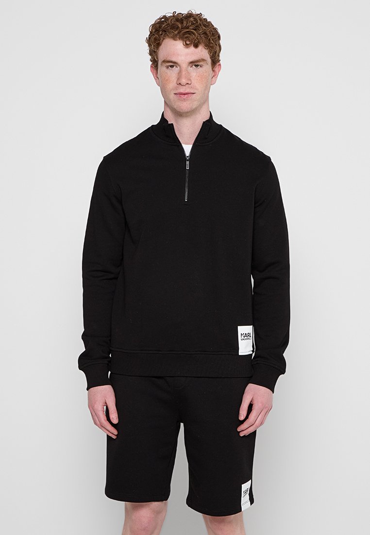 Schwarzer Fleece-Pullover mit einem halben Reißverschluss am Kragen, gerippten Bündchen und Saum. Kombiniert mit passender schwarzer Shorts mit einem Logopatch.