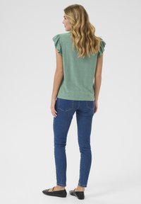 Top verde a maniche corte con maniche a volant, con dettagli in occhiello. Abbinato a jeans skinny blu e ballerine nere.